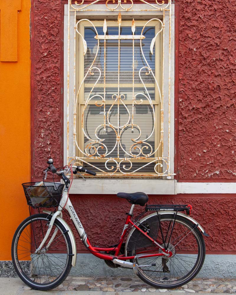 A red bike rests against a red wall with a white iron grill